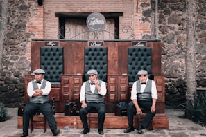 Three men in vests, bow ties, and hats seated on a wooden bench with three sections, each labeled with names Hugo, Paco, and Luis. The background consists of rustic brick and stone walls, and there is a wooden door behind them. The setting appears old-fashioned and reminiscent of early 20th-century establishments.