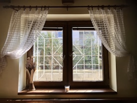 A wooden-framed window with sheer, patterned curtains on each side. Sunlight filters through the window, casting shadows on the interior wall. Outside, greenery and a metal grid structure are visible. On the window sill, there is a bundle of dried wheat tied together and a small white candle.