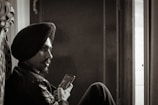 A sepia-toned photograph of a man with a turban, sitting and holding a smartphone, looking contemplatively towards a window with light streaming in.
