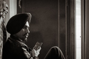 A sepia-toned photograph of a man with a turban, sitting and holding a smartphone, looking contemplatively towards a window with light streaming in.