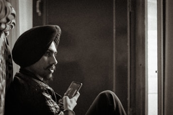 A sepia-toned photograph of a man with a turban, sitting and holding a smartphone, looking contemplatively towards a window with light streaming in.