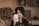 A person wearing a black turban and a black jacket is holding a smartphone. They are seated in a room with a bed covered in patterned sheets and a bookshelf filled with books in the background. A computer monitor is visible on a desk behind them.