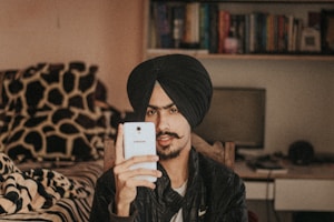 A person wearing a black turban and a black jacket is holding a smartphone. They are seated in a room with a bed covered in patterned sheets and a bookshelf filled with books in the background. A computer monitor is visible on a desk behind them.