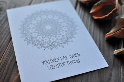 A white card with an intricate mandala design is placed on a wooden surface. The card bears an inspirational quote: 'YOU ONLY FAIL WHEN YOU STOP TRYING.' Several assorted seashells are scattered around the card.