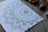 Intricate mandala pattern drawn on a piece of paper placed on a wooden surface with seashells nearby. The design includes floral and geometric elements.