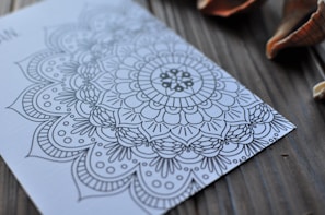 Intricate mandala pattern drawn on a piece of paper placed on a wooden surface with seashells nearby. The design includes floral and geometric elements.