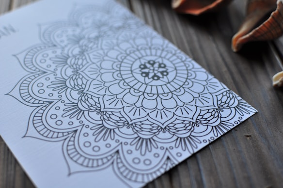 Intricate mandala pattern drawn on a piece of paper placed on a wooden surface with seashells nearby. The design includes floral and geometric elements.