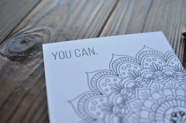 A close-up view of a card with intricate mandala artwork and the motivational text 'YOU CAN.' set on a wooden surface with visible grain patterns.