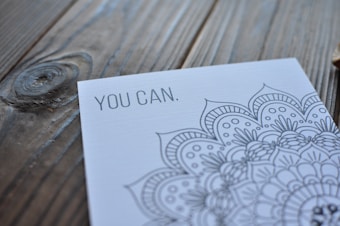 A close-up view of a card with intricate mandala artwork and the motivational text 'YOU CAN.' set on a wooden surface with visible grain patterns.