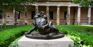 A bronze sculpture depicting a muscular figure wrestling a lion, set on a stone pedestal. The sculpture is surrounded by manicured greenery with a historic building featuring stone columns and arches in the background.