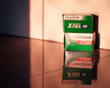 Two boxes of Fujifilm camera film sit on a reflective surface, with warm, soft lighting casting shadows and highlights. The top box is labeled 'FUJIFILM Superia X-TRA 400', and the bottom one is marked 'FUJICOLOR C200'. The labels are clearly visible, and the reflection enhances the warm tones.