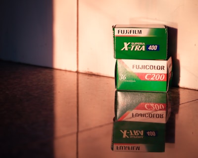 Two boxes of Fujifilm camera film sit on a reflective surface, with warm, soft lighting casting shadows and highlights. The top box is labeled 'FUJIFILM Superia X-TRA 400', and the bottom one is marked 'FUJICOLOR C200'. The labels are clearly visible, and the reflection enhances the warm tones.