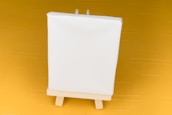 A small blank white canvas stands on a wooden easel, set against a mustard yellow background.