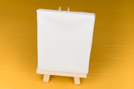 A small blank white canvas stands on a wooden easel, set against a mustard yellow background.