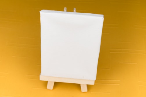 A small blank white canvas stands on a wooden easel, set against a mustard yellow background.