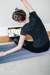 A woman is performing a seated side stretch on a yoga mat indoors, with her back facing the camera. Her hair is styled in two small buns, and she is wearing a dark t-shirt and leggings. A laptop displaying a yoga video is placed in front of her on the floor.