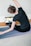A woman is performing a seated side stretch on a yoga mat indoors, with her back facing the camera. Her hair is styled in two small buns, and she is wearing a dark t-shirt and leggings. A laptop displaying a yoga video is placed in front of her on the floor.