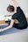 A woman is performing a seated side stretch on a yoga mat indoors, with her back facing the camera. Her hair is styled in two small buns, and she is wearing a dark t-shirt and leggings. A laptop displaying a yoga video is placed in front of her on the floor.