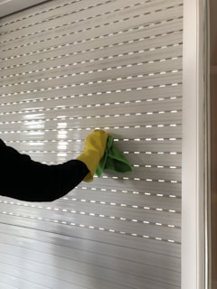 A person wearing a yellow glove is cleaning a white shutter with a green cloth. The shutter has horizontal slats and some light peeks through.