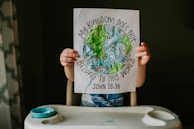 A child holding up a piece of paper with a colorful and textured drawing of Earth surrounded by flowers. The text around the drawing reads 'My Kingdom Does Not Belong to This World' with the biblical reference 'John 18:36'.