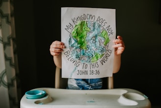 A child holding up a piece of paper with a colorful and textured drawing of Earth surrounded by flowers. The text around the drawing reads 'My Kingdom Does Not Belong to This World' with the biblical reference 'John 18:36'.