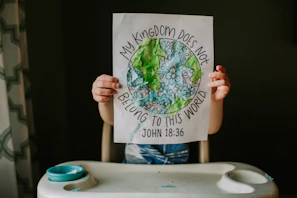 A joyful child holding up a completed coloring activity featuring a Bible verse