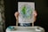 A child holding up a piece of paper with a colorful and textured drawing of Earth surrounded by flowers. The text around the drawing reads 'My Kingdom Does Not Belong to This World' with the biblical reference 'John 18:36'.
