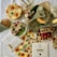 A variety of items are displayed on a table covered with a white cloth. There are food items such as bread, cucumbers, greens, pumpkin, and cherry tomatoes wrapped or covered in colorful cloths with different patterns. These cloths include sunflower prints, leaf motifs, butterfly designs, and floral patterns. Two cylindrical packages wrapped in patterned paper, a cutting board, and a document related to the product or recipe are also present.