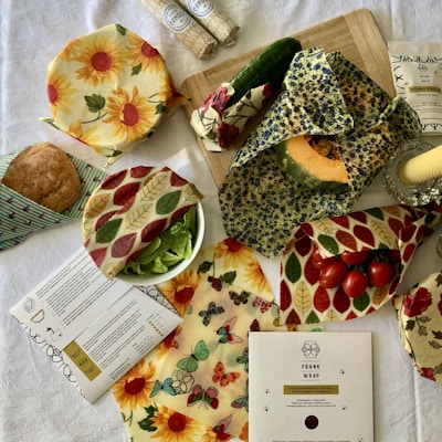 A variety of items are displayed on a table covered with a white cloth. There are food items such as bread, cucumbers, greens, pumpkin, and cherry tomatoes wrapped or covered in colorful cloths with different patterns. These cloths include sunflower prints, leaf motifs, butterfly designs, and floral patterns. Two cylindrical packages wrapped in patterned paper, a cutting board, and a document related to the product or recipe are also present.