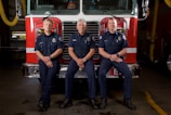 Three firefighters are in uniform, sitting and standing in front of a fire truck. They are inside a fire station, and the truck features a red front with chrome details.