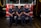 Three firefighters are in uniform, sitting and standing in front of a fire truck. They are inside a fire station, and the truck features a red front with chrome details.
