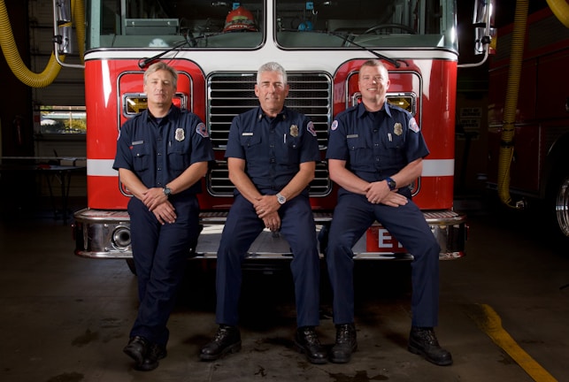 Three firefighters are in uniform, sitting and standing in front of a fire truck. They are inside a fire station, and the truck features a red front with chrome details.