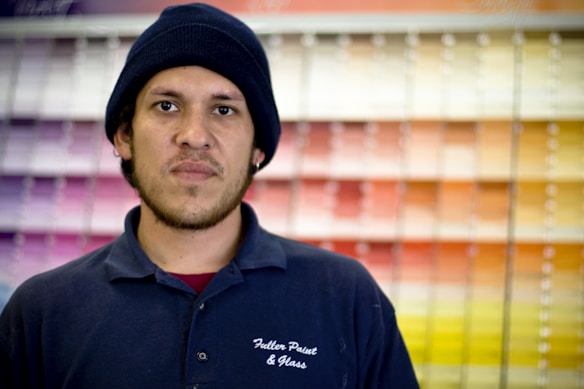 A man wearing a dark beanie and a navy blue polo shirt stands in front of a display of colorful paint swatches. The shirt has a logo that reads 'Fuller Paint & Glass'. The paint swatches are arranged in a gradient, creating a vibrant backdrop.