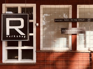 A wooden building facade with white-framed windows and a large black sign featuring a prominent letter 'R' and the word 'workshop' below. Additional rectangular signs read 'FINNISH ROCK TO THE WORLD' with a small hand gesture and 'rockersworkshop.com'. A wire mesh grid covers the windows.