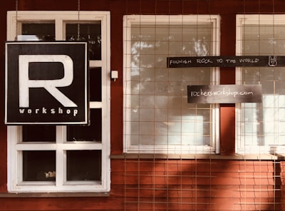 A wooden building facade with white-framed windows and a large black sign featuring a prominent letter 'R' and the word 'workshop' below. Additional rectangular signs read 'FINNISH ROCK TO THE WORLD' with a small hand gesture and 'rockersworkshop.com'. A wire mesh grid covers the windows.