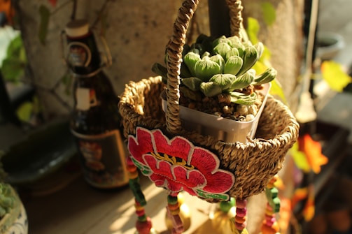 A woven basket hangs with an intricate embroidered flower design on the front. Inside the basket sits a small pot containing a vibrant succulent plant. The scene is warmly lit, casting soft shadows, and features a decorative bottle in the background.