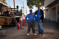 Two men in work attire stand confidently in front of construction equipment. The setting appears to be an outdoor worksite with an asphalt ground and surrounding safety cones. Utility poles and a palm tree are visible in the background against a clear sky.