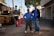Two men in work attire stand confidently in front of construction equipment. The setting appears to be an outdoor worksite with an asphalt ground and surrounding safety cones. Utility poles and a palm tree are visible in the background against a clear sky.