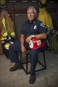 A firefighter in uniform sits on a chair in a room with various firefighting gear. The individual is holding a red firefighter helmet labeled with 'Captain' and 'E1'. The background includes hanging jackets and helmets, and the firefighter’s expression is serious and composed.