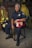 A firefighter in uniform sits on a chair in a room with various firefighting gear. The individual is holding a red firefighter helmet labeled with 'Captain' and 'E1'. The background includes hanging jackets and helmets, and the firefighter’s expression is serious and composed.