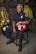 A firefighter in uniform sits on a chair in a room with various firefighting gear. The individual is holding a red firefighter helmet labeled with 'Captain' and 'E1'. The background includes hanging jackets and helmets, and the firefighter’s expression is serious and composed.