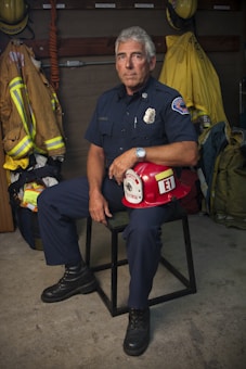 A firefighter in uniform sits on a chair in a room with various firefighting gear. The individual is holding a red firefighter helmet labeled with 'Captain' and 'E1'. The background includes hanging jackets and helmets, and the firefighter’s expression is serious and composed.