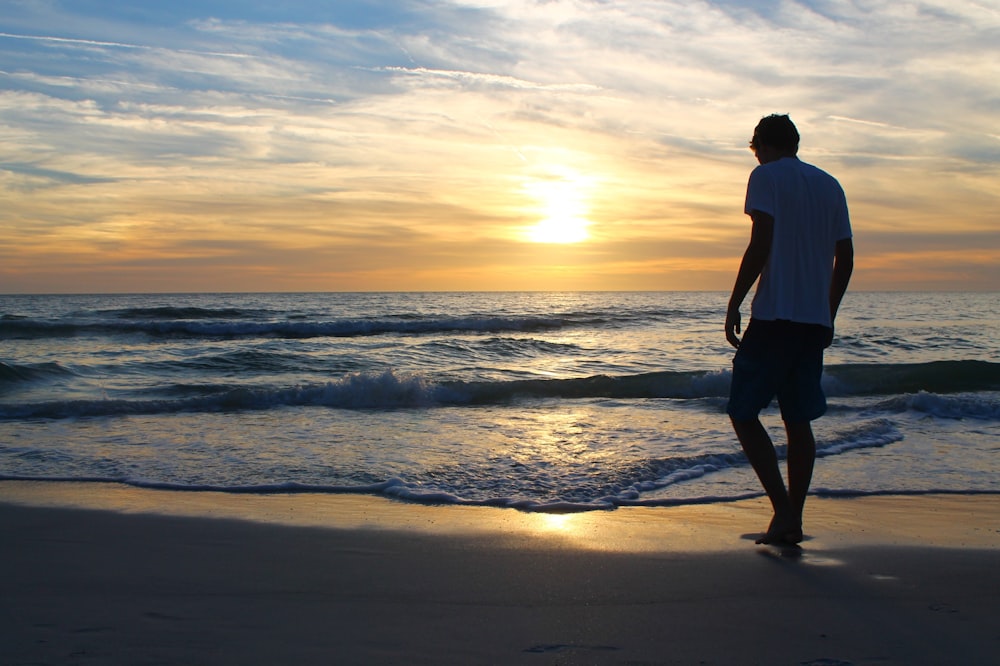 man in white t-shirt and black shorts standing on seashore during sunset