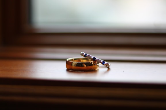 A close-up of a gold ring accompanied by a delicate bracelet adorned with small beads, resting on a wooden surface near a window. The background is softly blurred, creating a sense of calm and focus on the jewelry.