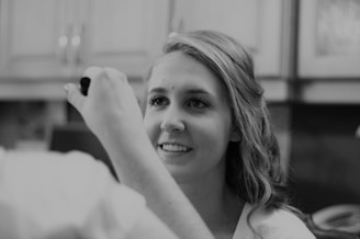 A person with long hair is smiling while looking upwards. Another person is holding a makeup brush near the face of the smiling person. The setting appears to be a room with light-colored cabinets in the background, suggesting a home or casual environment.