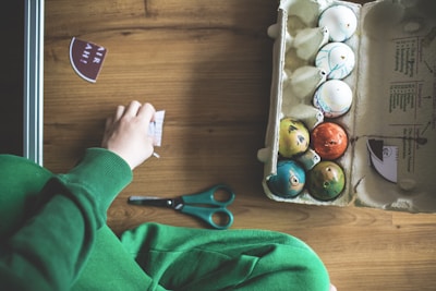 A person is seated at a wooden table, engaged in a craft activity involving colorful, decorated eggs placed in an open egg carton. Nearby, a pair of green scissors lies on the table. The person is wearing a green sleeve, and their hand is focused on manipulating a small piece of printed material.