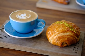 Close-up of a steaming cup of coffee with latte art on top, placed next to a croissant.