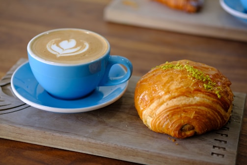 Close-up of a steaming cup of coffee with latte art on top, placed next to a croissant.