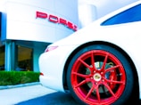 A sleek white sports car with vibrant red wheels, parked in front of a luxury car dealership. The dealership building is modern with a minimalist design, and the brand logo is prominently displayed in bold red letters.