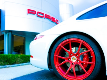 A sleek white sports car with vibrant red wheels, parked in front of a luxury car dealership. The dealership building is modern with a minimalist design, and the brand logo is prominently displayed in bold red letters.
